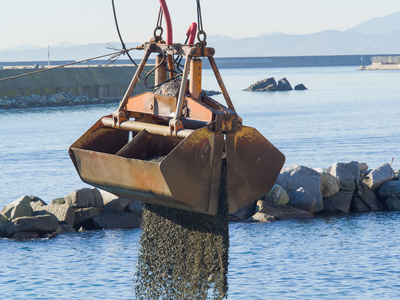 Dredge Clamshell Bucket unloading gravel in the water of a port next to the shore to replenish a beach